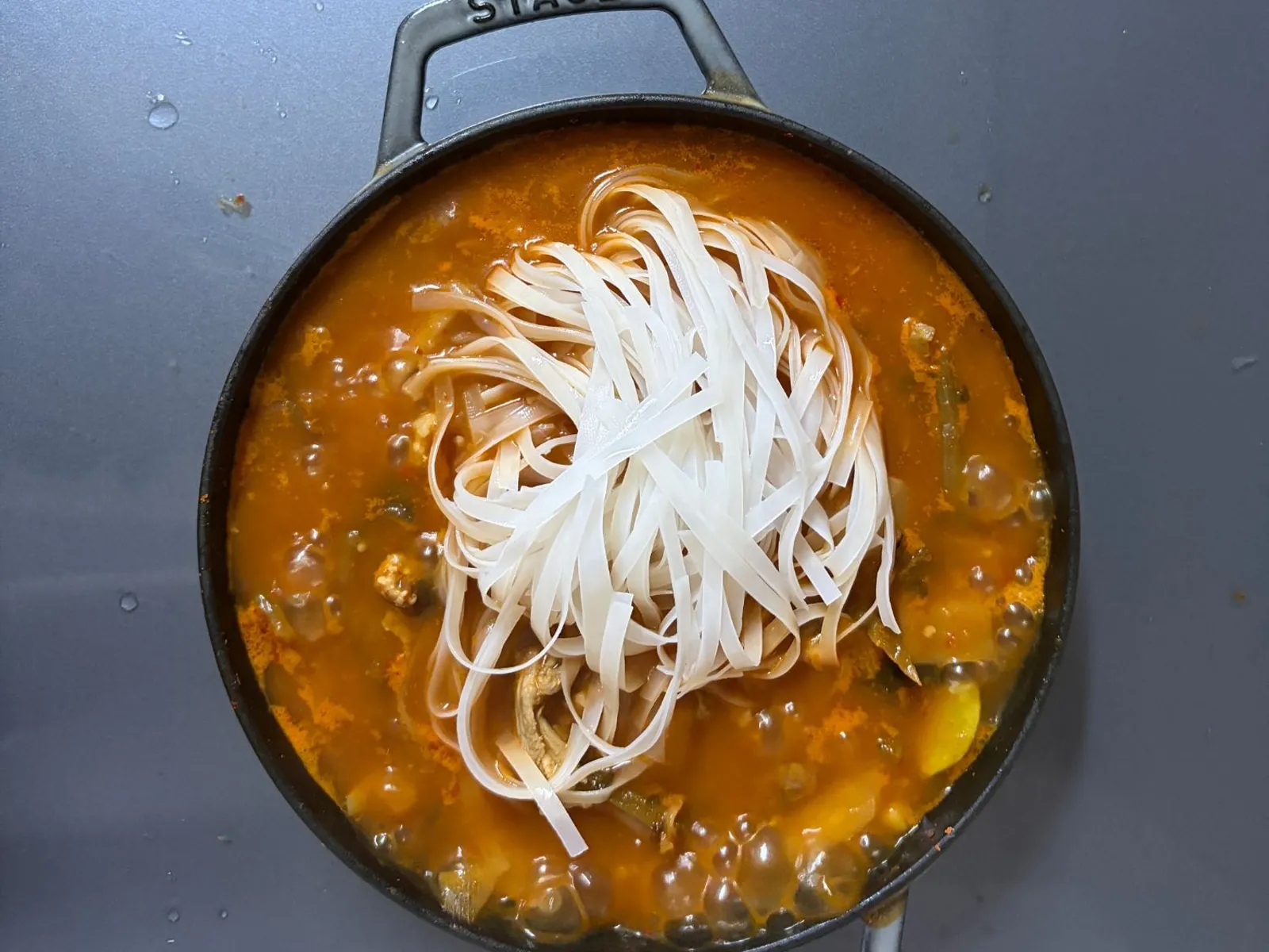 Soaked flat rice noodles placed on top of simmering kimchi jjigae in a cast iron pan