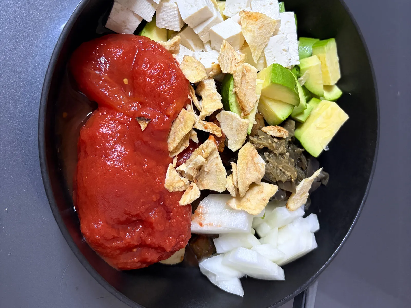 Canned whole tomatoes, cubed tofu, zucchini, onion, eggplant kimchi and dried apple slices in a black cast iron pan before cooking