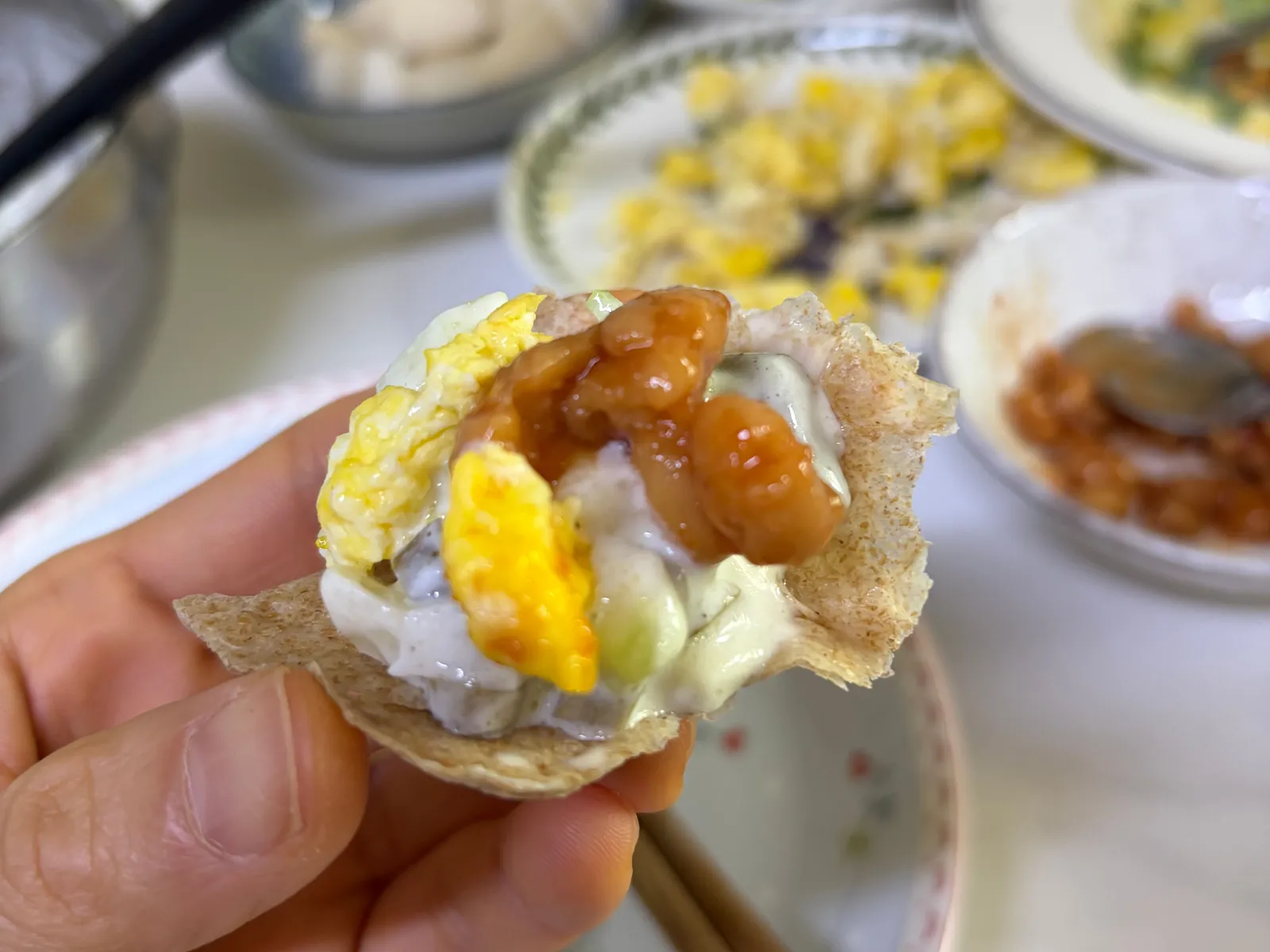 A torn piece of tortilla topped with oiji tzatziki, scrambled egg and baked beans, held in one hand over a breakfast table
