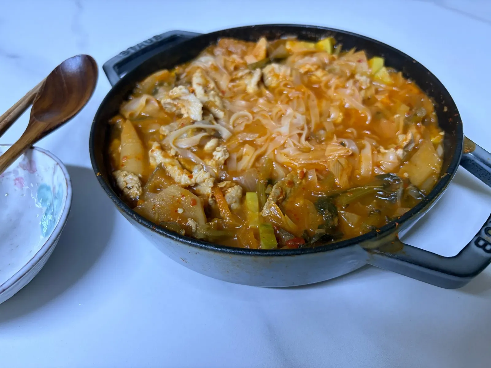 Kimchi jjigae with flat rice noodles, pork and zucchini in a cast iron pan on a white table
